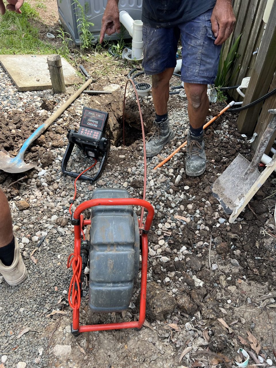 Person uses CCTV Pipe Inspection camera on underground pipe by a freshly dug hole, tools nearby.