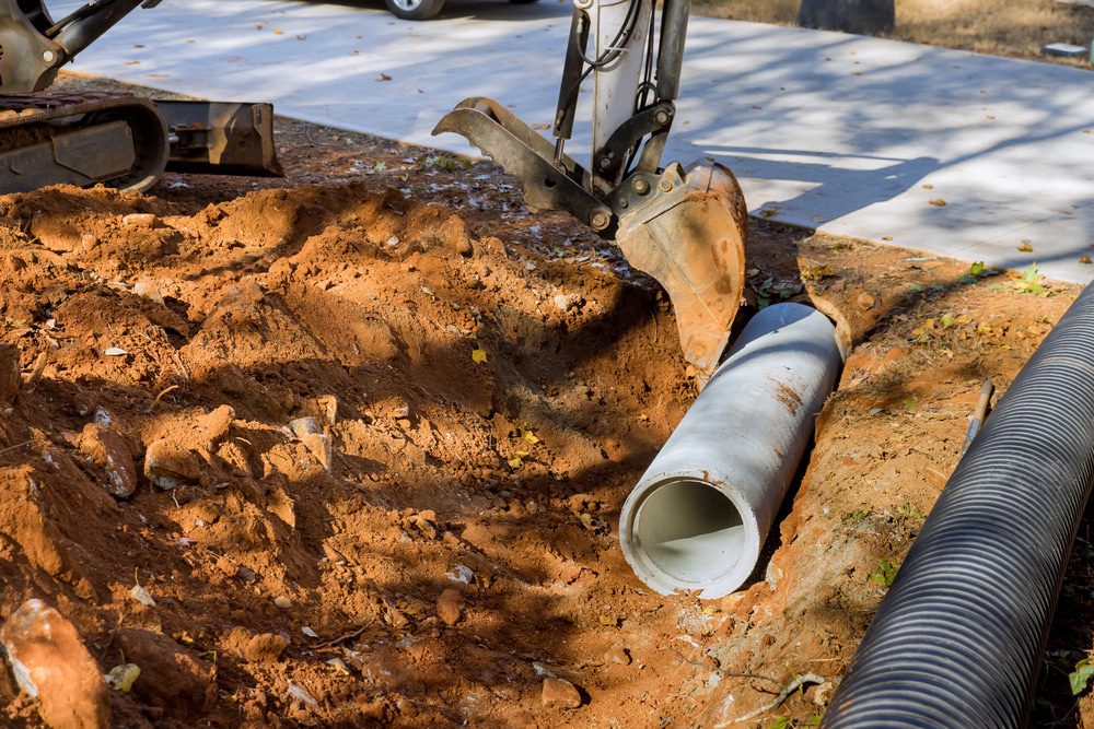 Construction excavator bucket over a concrete pipe in a trench beside a corrugated drainage pipe.