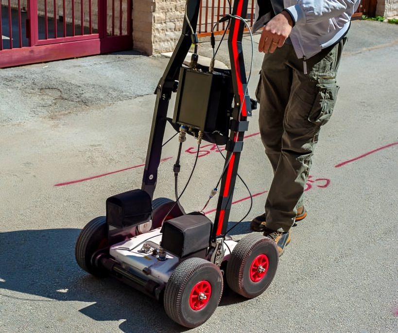 Person using Ground Penetrating Radar (GPR) on a paved surface with red spray paint for subsurface investigations.