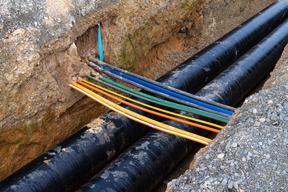 Two large black pipes run parallel in a trench with underground electrical cables crossing above, exposed dirt visible.