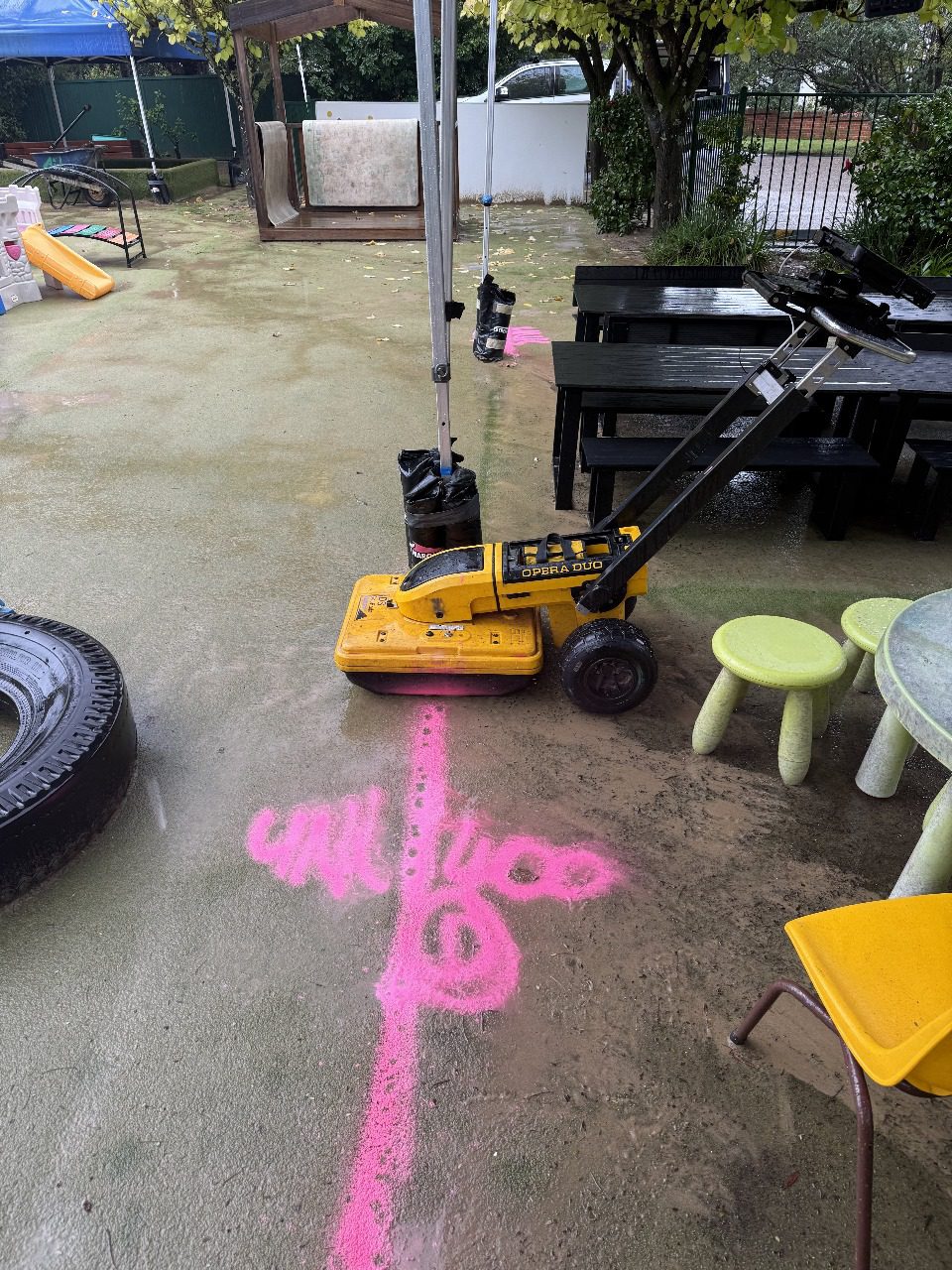 yellow concrete scanning radar cart on wet ground near pink line, children’s tables and chairs in background.